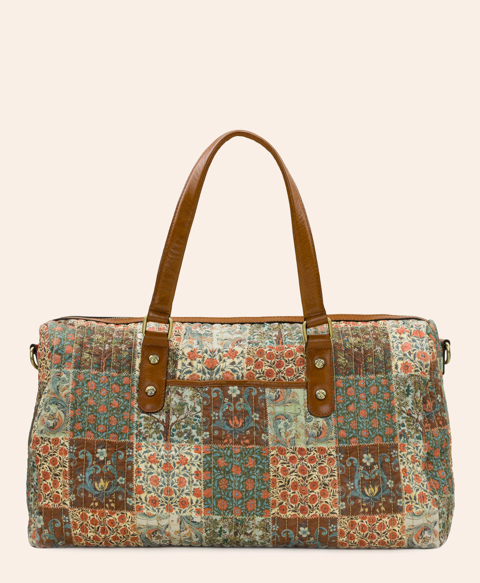 A patchwork quilted duffel bag with a multicolor floral and paisley pattern, featuring brown faux-leather handles and gold-tone hardware, set against a solid light beige background.