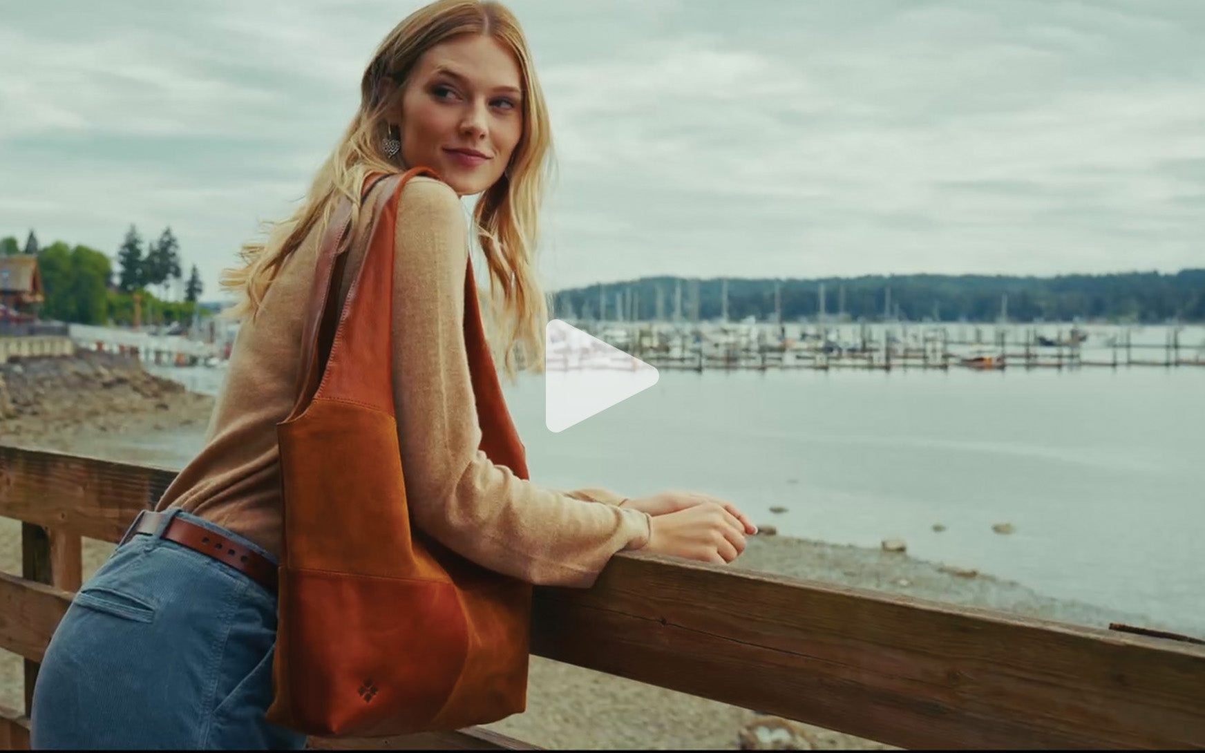 A woman with blonde hair leans on a wooden railing by the water, wearing a beige sweater, blue jeans, and carrying a large brown leather tote bag. A white play icon is overlaid in the center of the image.