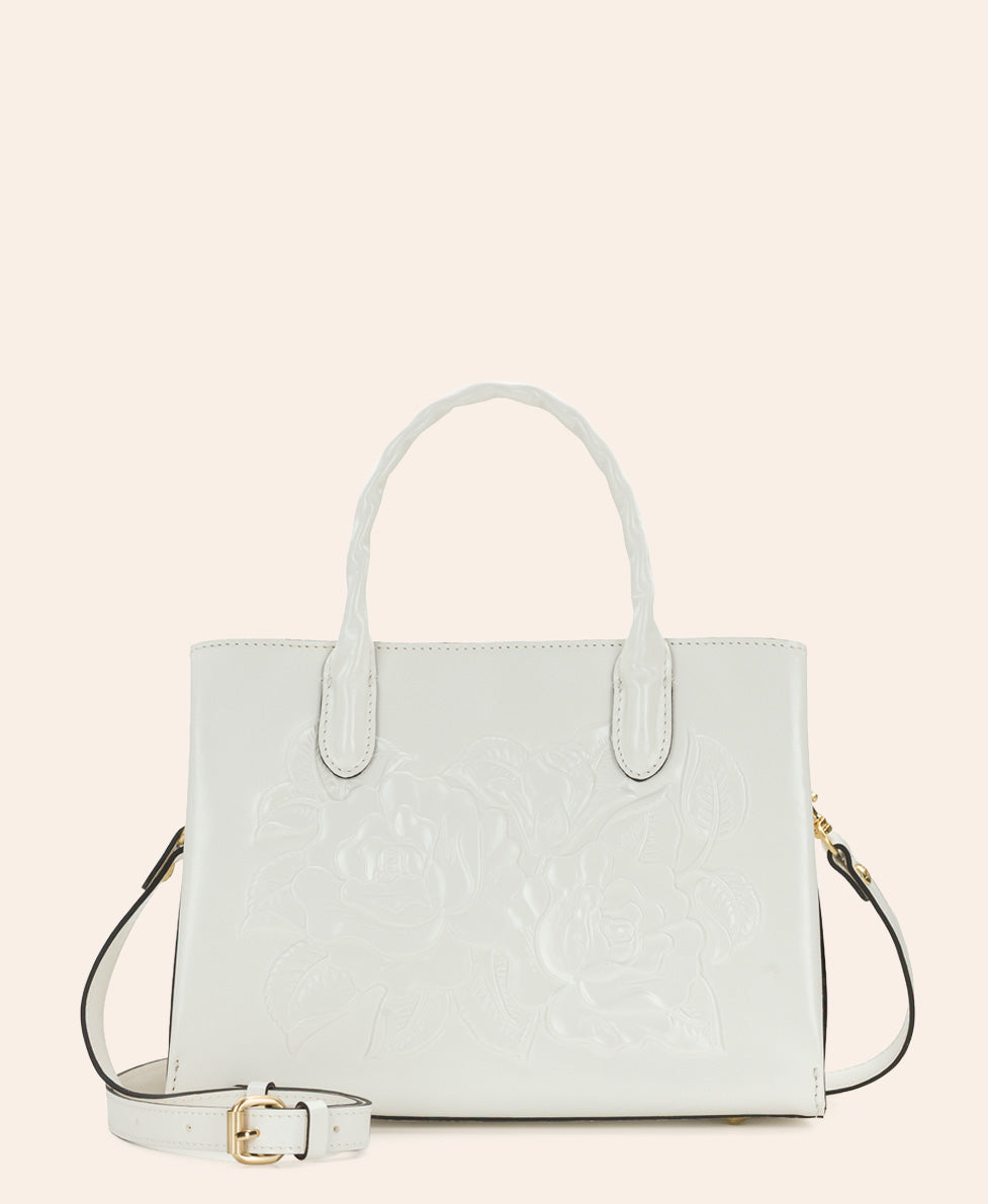A light off-white leather handbag with embossed floral detailing, featuring dual top handles and a matching detachable shoulder strap with a gold buckle, set against a solid cream background.