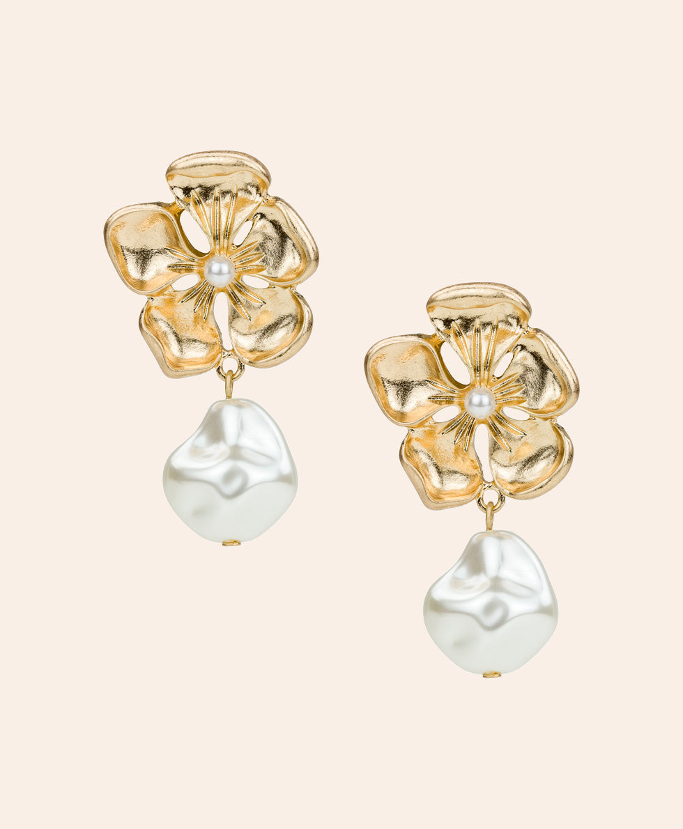 A pair of gold flower-shaped dangle earrings with a small white pearl at the center and a larger baroque-style white pearl hanging below, shown on a light beige background.
