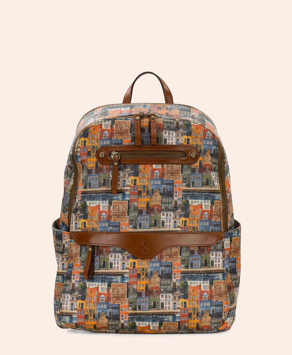 A backpack featuring a colorful, dense architectural illustration pattern of buildings in various shades of blue, orange, yellow, and red, accented with brown leather trim, a front zip pocket, and a top handle, set against a solid light beige background.