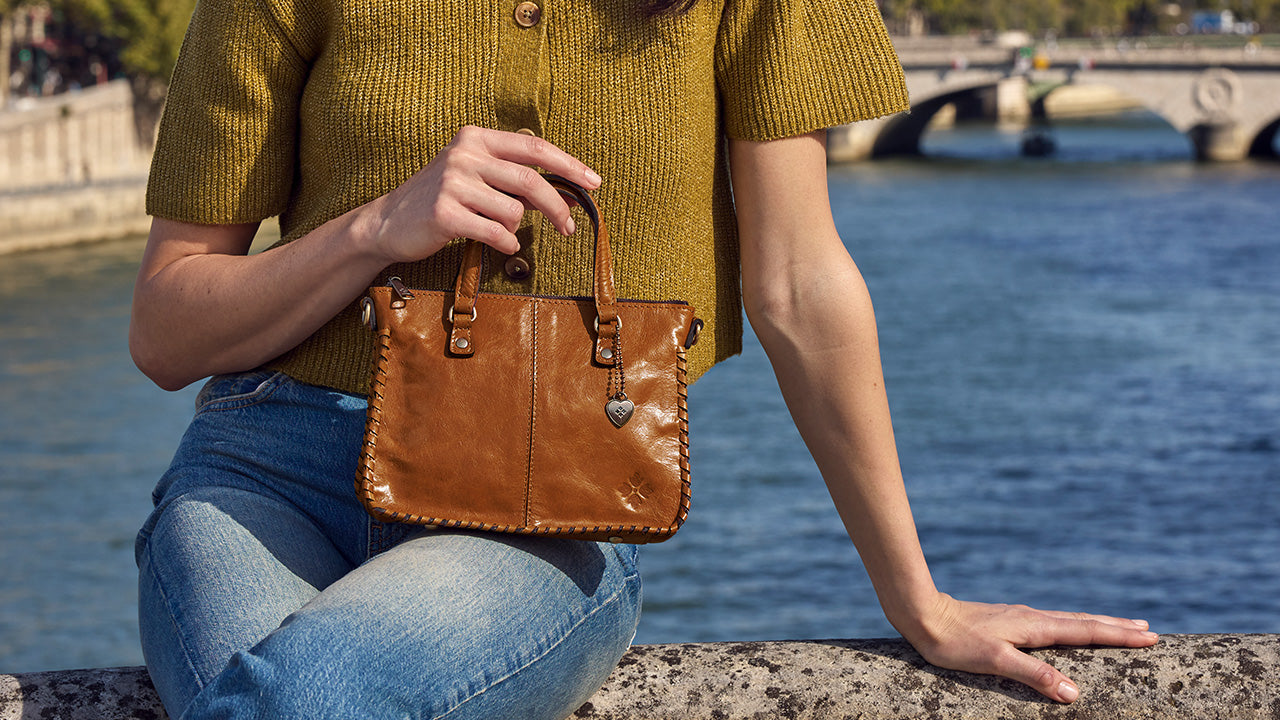 A woman wearing a mustard yellow knit top and blue jeans sits by a river, holding a brown leather handbag with brass hardware.