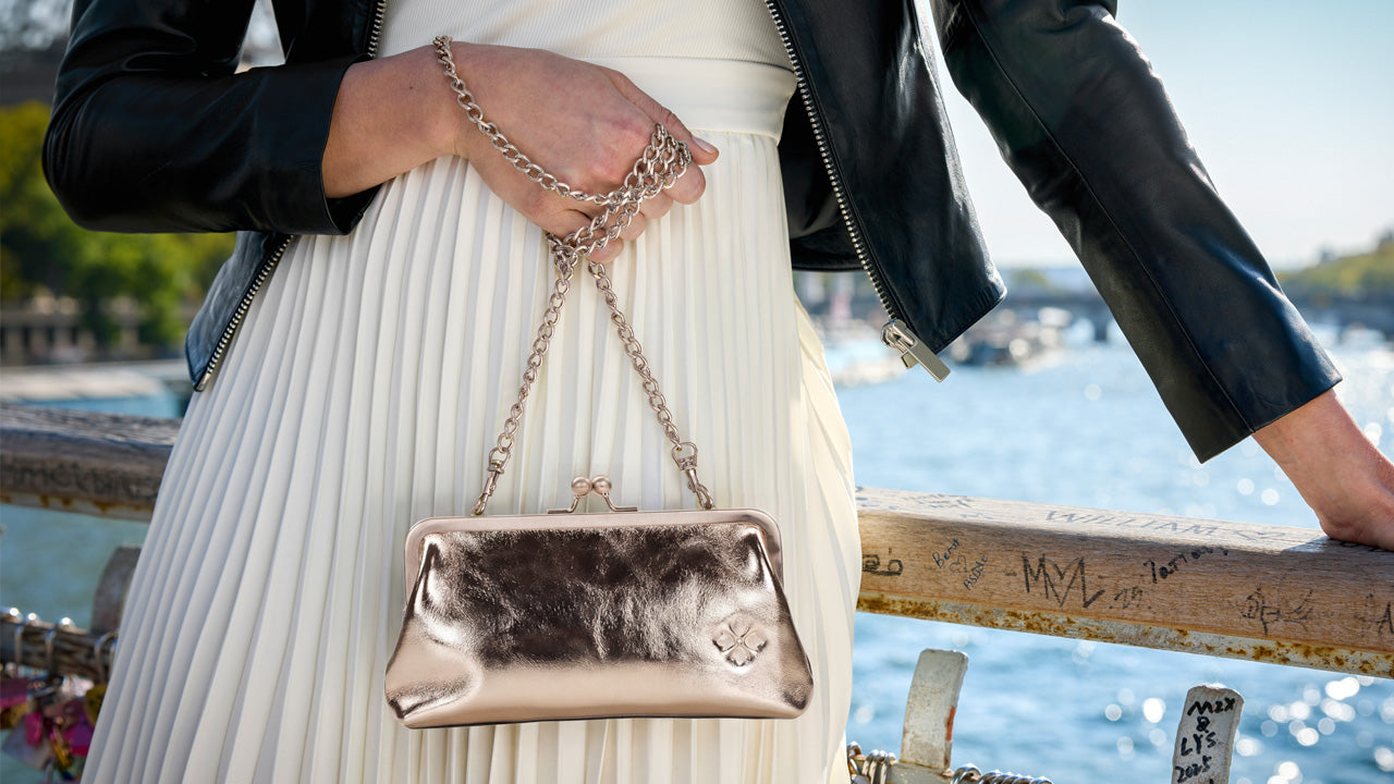 A person in a white pleated dress and black leather jacket holds a metallic champagne clutch bag with a chain strap, set against a blurred outdoor waterfront background.