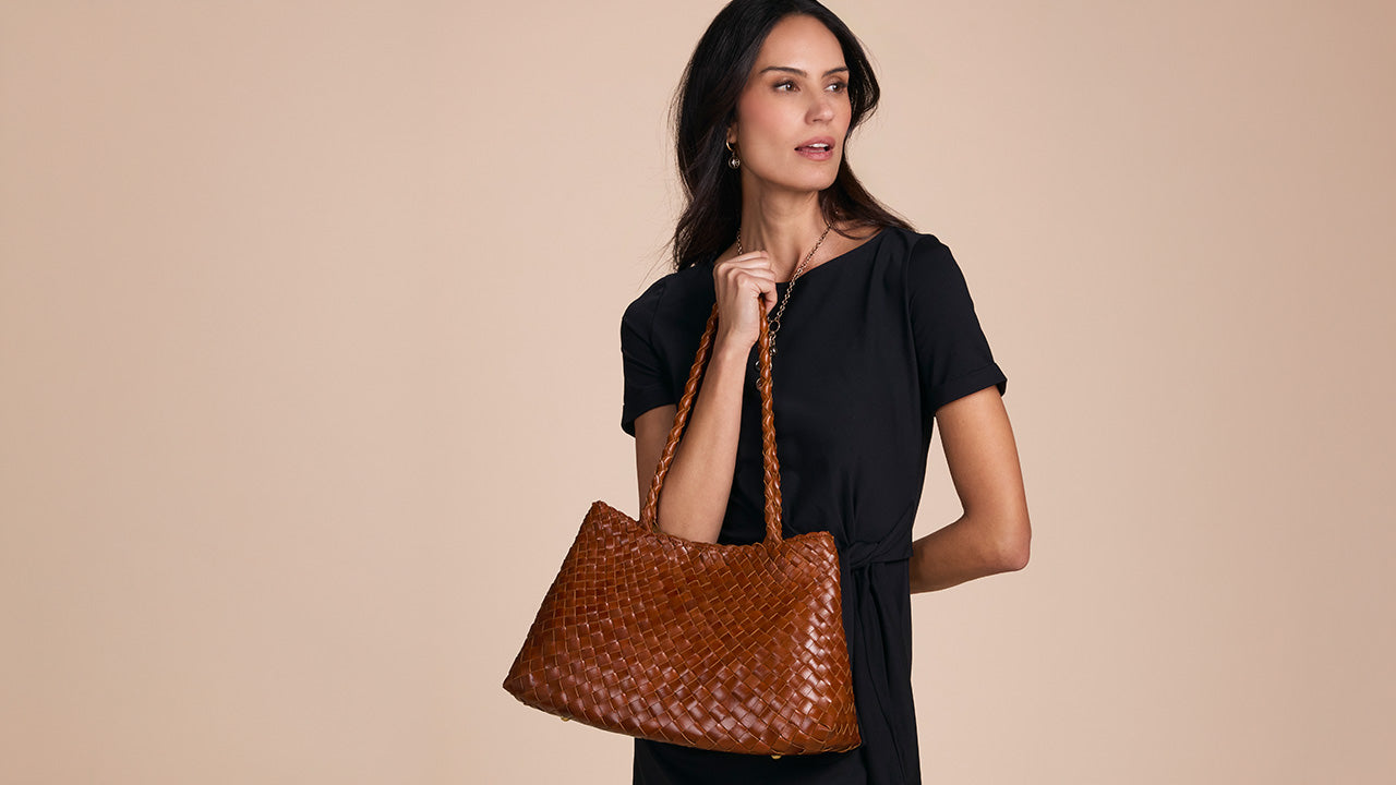 A woman wearing a black dress holds a large brown woven leather tote bag against a solid beige background.