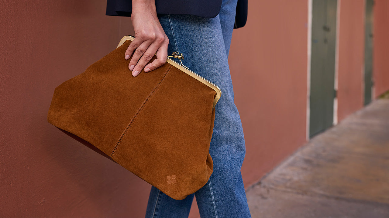 A person wearing blue jeans holds a large brown suede clutch bag with a gold-tone metal frame against a terracotta-colored wall.