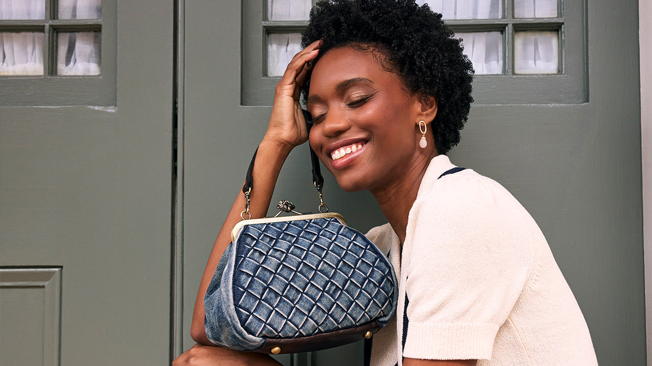 A smiling woman with short black curly hair holds a blue quilted handbag with gold-tone hardware in front of a gray door.