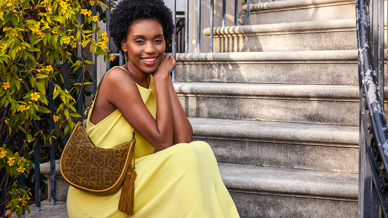 A woman with curly black hair sits on outdoor stone steps wearing a bright yellow dress and carrying a brown monogram shoulder bag with a tassel.