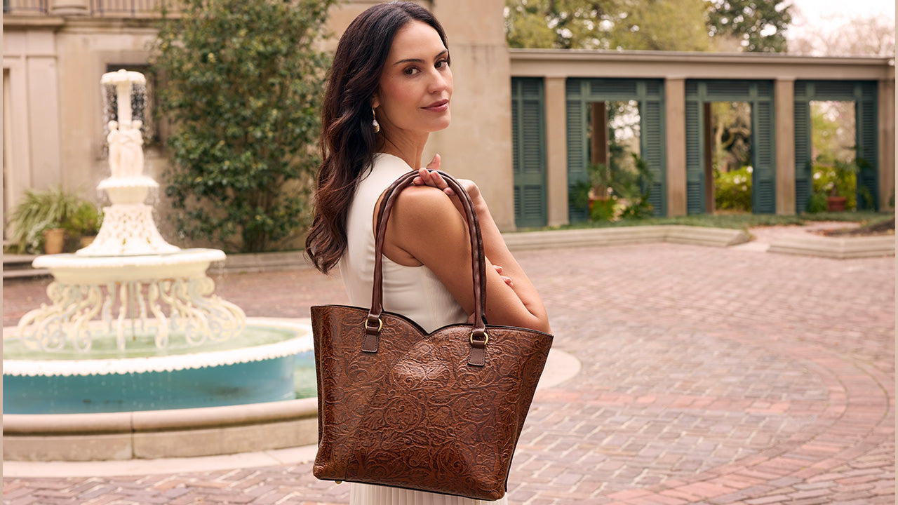 A woman in a white sleeveless top carries a large brown embossed leather tote bag with double shoulder straps, standing in a courtyard with a fountain and building in the background.