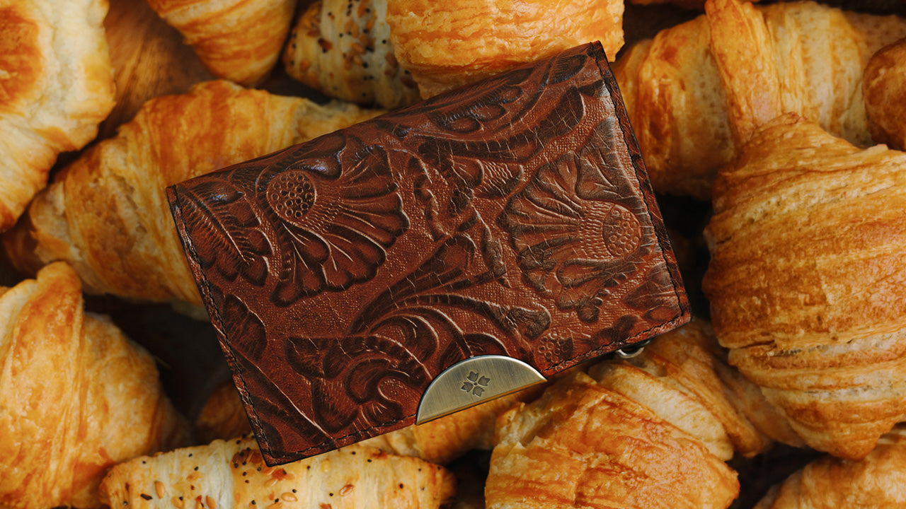 A dark brown embossed leather card holder wallet with a silver metal snap button, resting on a pile of golden-brown croissants.