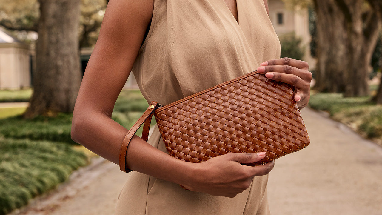 A woman in a beige dress holds a brown woven leather clutch bag with a wrist strap in an outdoor park setting.