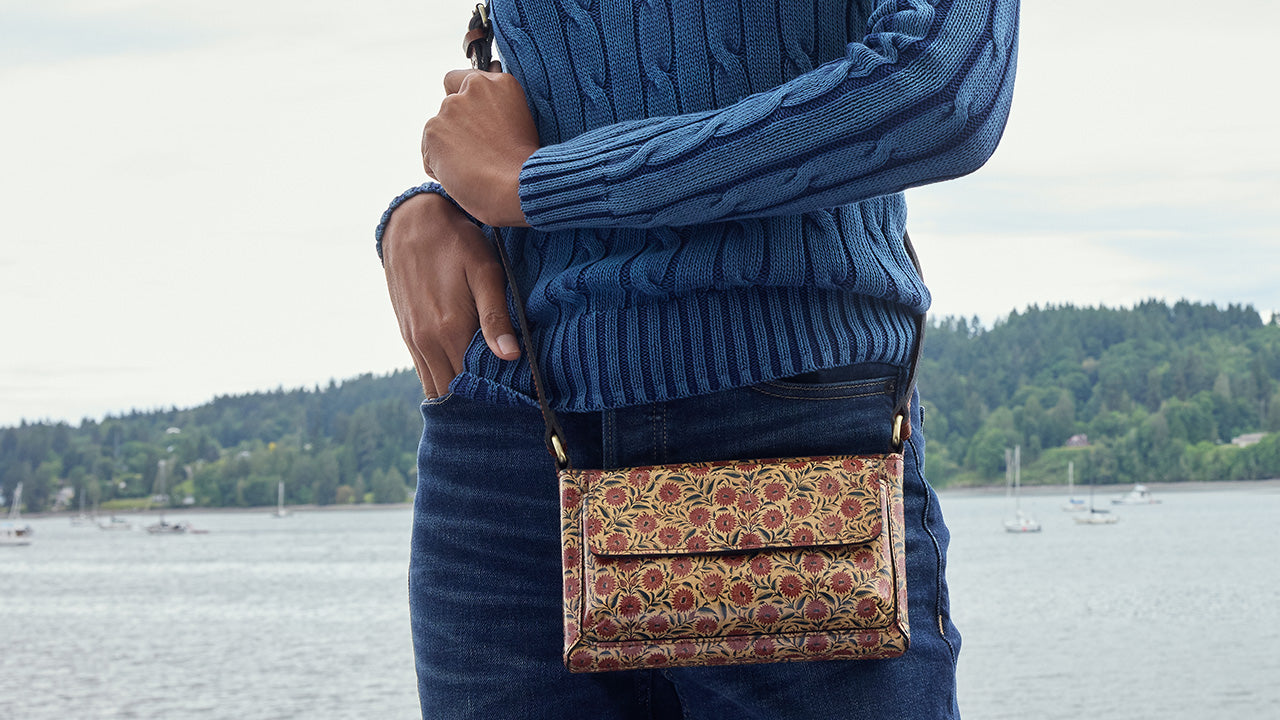 A person wearing a blue cable-knit sweater and dark jeans carries a small rectangular crossbody bag with a brown, gold, and red floral pattern. The background shows a body of water and a tree-lined shore under an overcast sky.