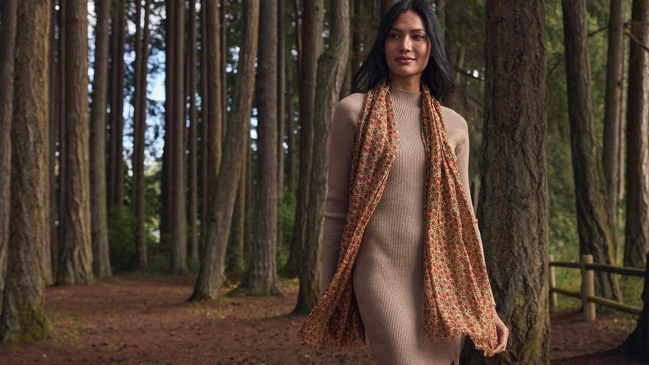 A woman stands in a forest wearing a beige knit dress and a rust-orange patterned scarf.