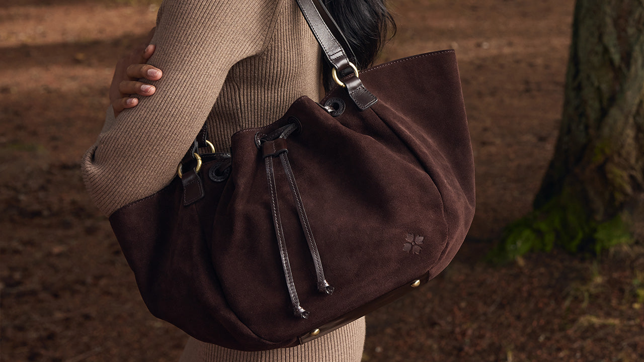 A person wearing a beige ribbed sweater carries a large dark brown suede drawstring tote bag outdoors near a tree trunk.