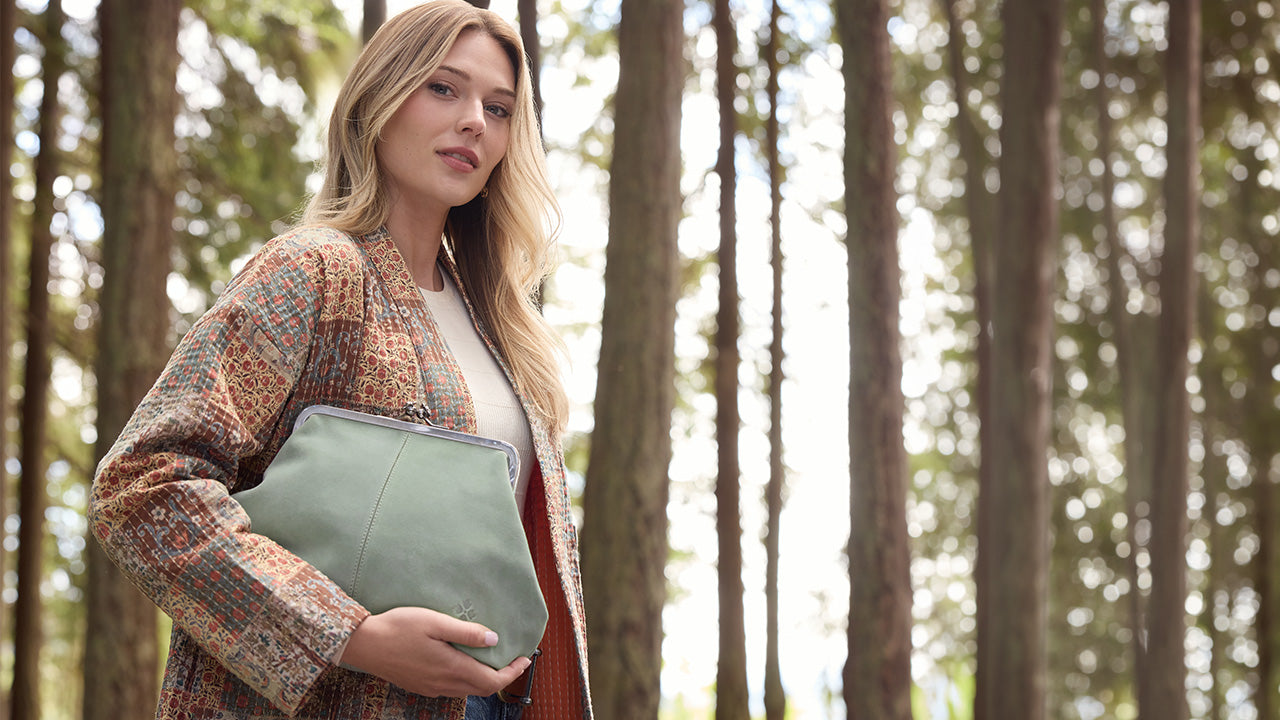 A woman standing in a forest holding a sage green leather clutch bag.