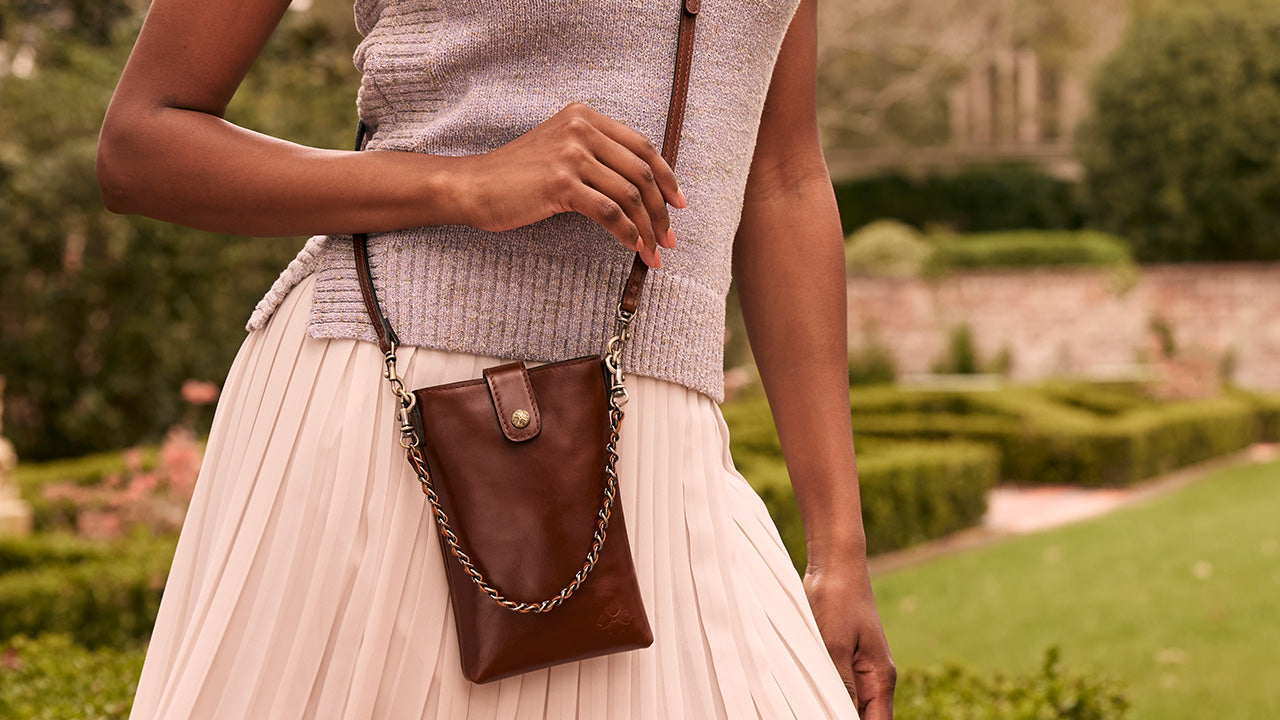 A woman wearing a light gray knit top and a cream pleated skirt carries a small brown leather crossbody bag with a gold-tone chain strap in an outdoor garden setting.