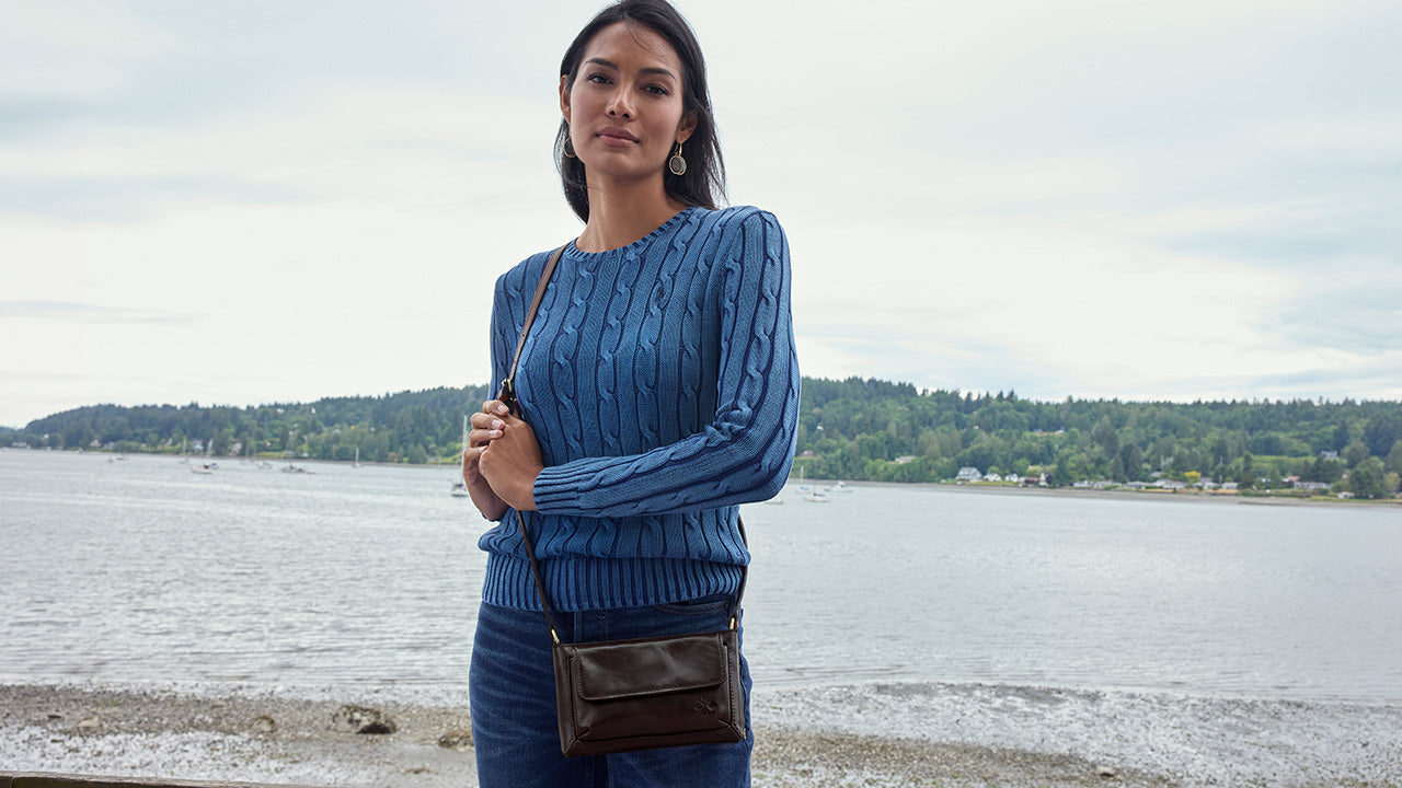 A woman stands outdoors by the water wearing a blue cable-knit sweater and dark blue jeans, with a brown crossbody bag. A shoreline with trees and an overcast sky are visible in the background.