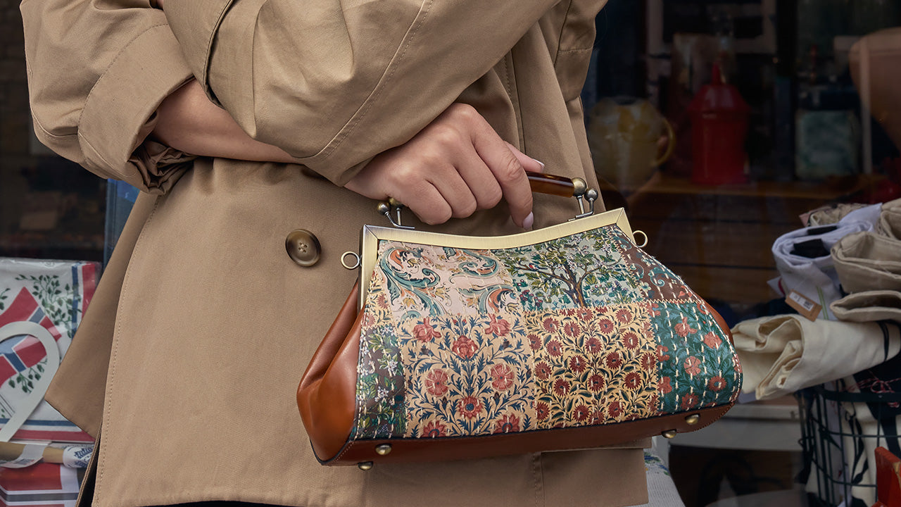A person in a beige trench coat holds a vintage-style handbag with a floral brocade pattern and brown leather trim in an outdoor market setting.