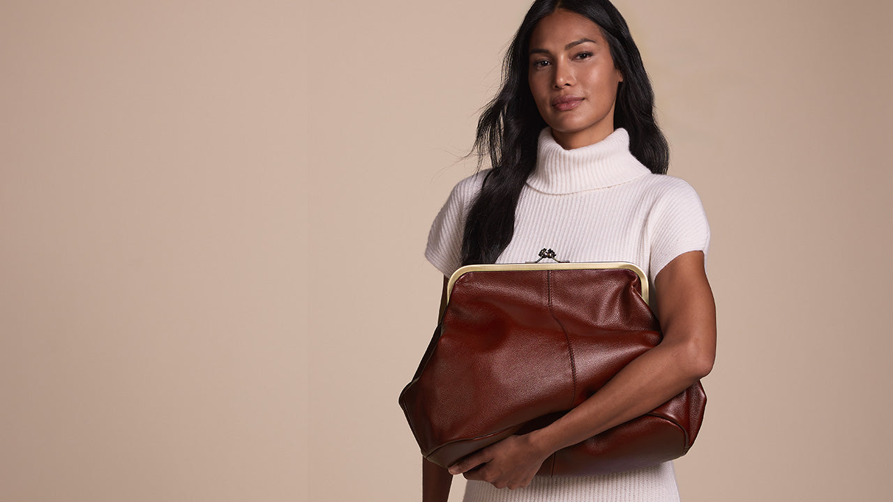 A woman wearing a white turtleneck sweater holds a large brown leather shoulder bag with a gold-tone frame clasp against a solid beige background.