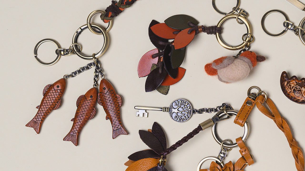 A styled flat-lay of several leather keychains on a light beige background. The keychains include fish-shaped leather charms, a key-shaped charm with a silver-toned ornate key, a small bird charm, and brown leather straps.