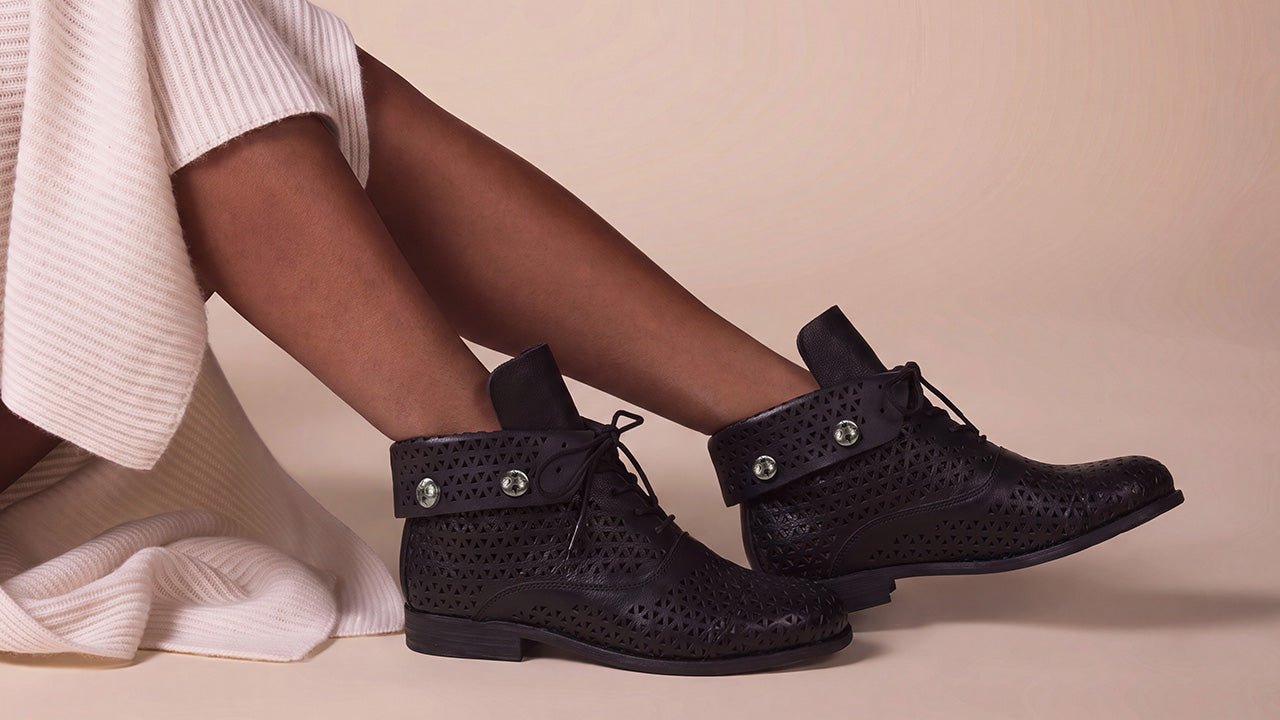 A person seated with legs crossed wearing black perforated ankle boots with silver-tone hardware and white ribbed knit socks, against a warm beige studio background.