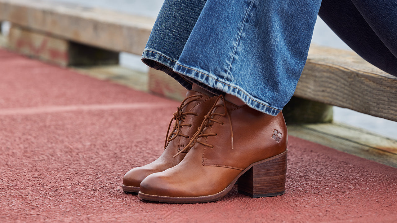 A close-up of a person wearing brown leather lace-up ankle boots with a block heel, paired with blue jeans, sitting on a wooden bench on a red textured surface.