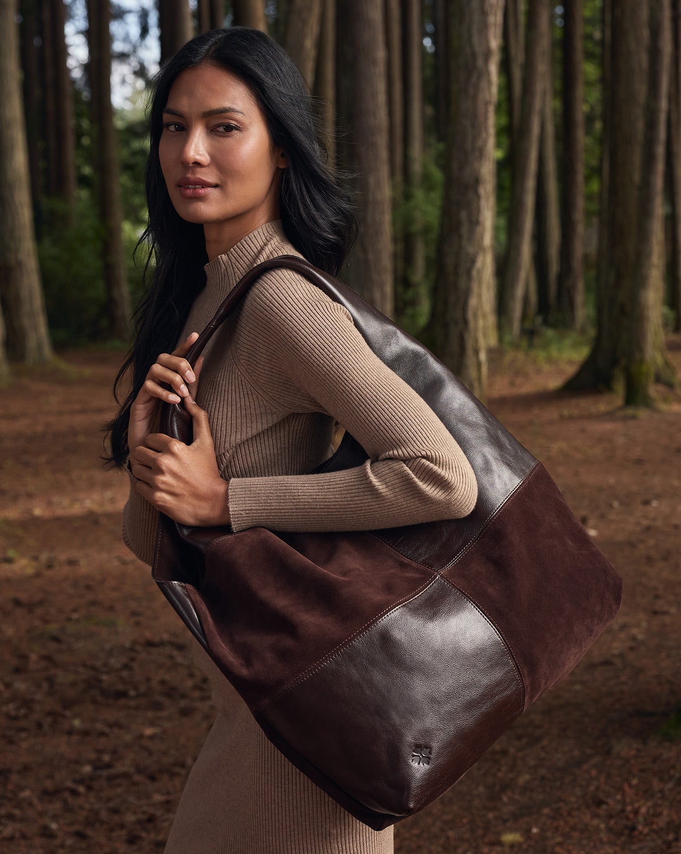 A woman in a beige ribbed turtleneck sweater stands in a forest holding a large brown leather and suede tote bag.
