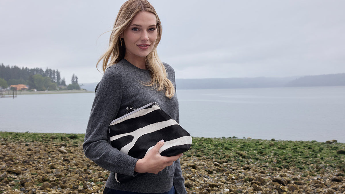A woman standing on a rocky shoreline holding a black and white zebra-patterned clutch bag, with a body of water and distant land in the background.