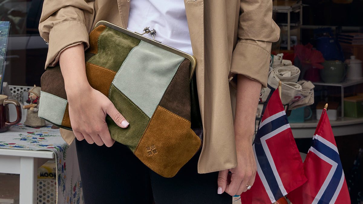 A person wearing a beige trench coat and white top holds a square patchwork clutch bag made of muted green, brown, and tan suede-like panels. The scene appears to be a shop interior with shelves and flags in the background.