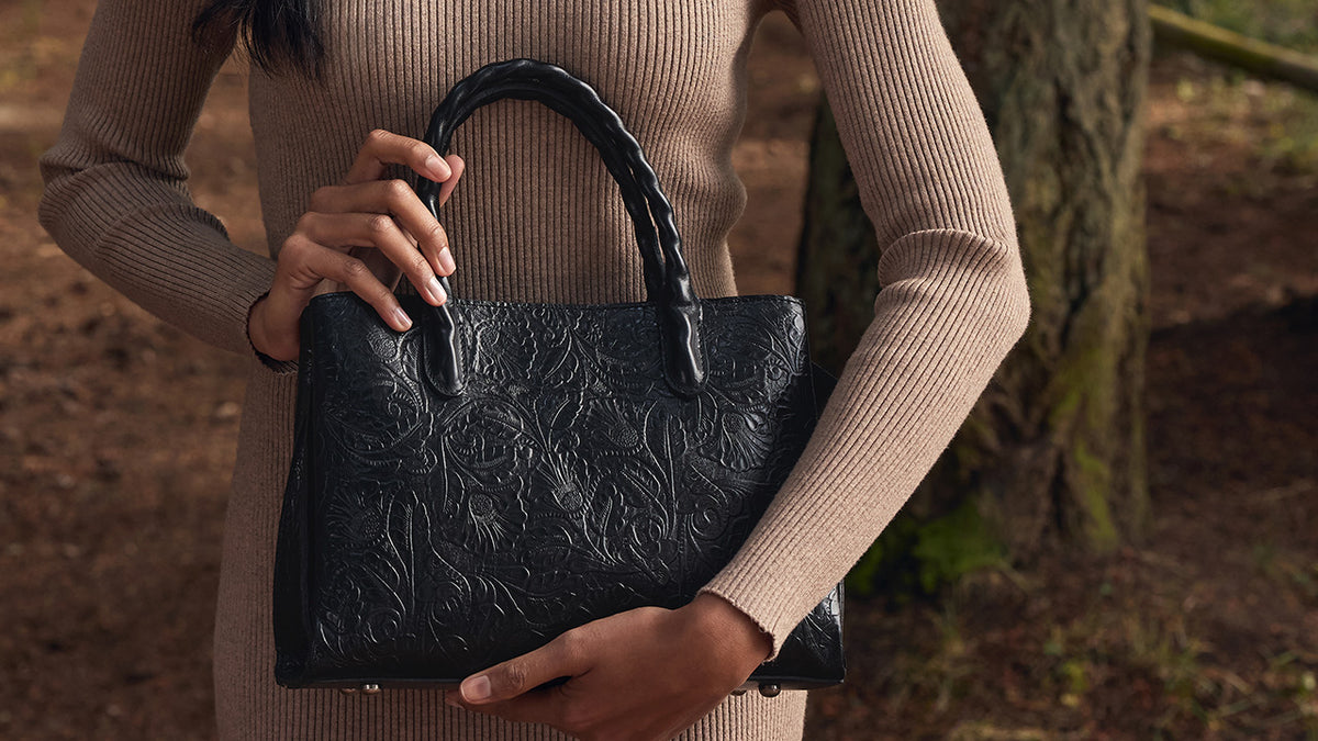 A person in a beige ribbed sweater holds a black embossed leather handbag with a braided top handle in an outdoor forest setting.
