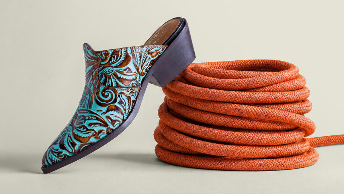 A single pointed-toe mule boot with turquoise, brown, and black tooled leather detailing, posed next to a coiled orange rope on a light beige background.