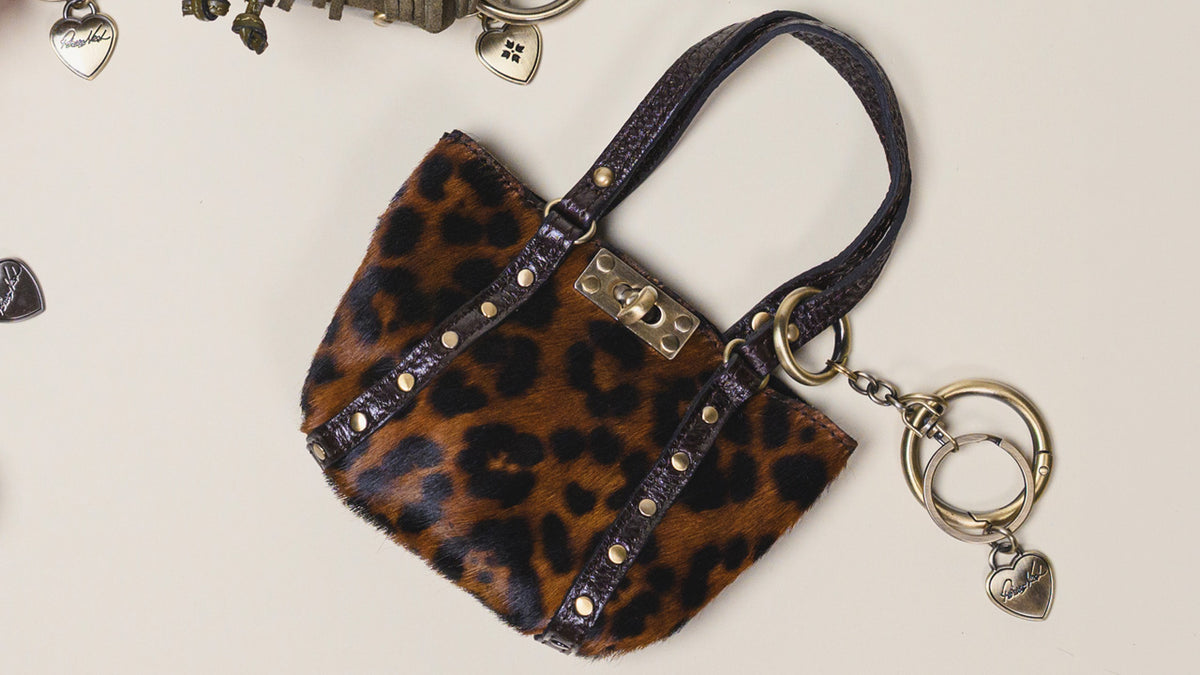 A small leopard-print handbag with dark purple leather trim and gold-tone hardware, including a turn-lock clasp and a key ring charm with heart-shaped tags, photographed on a light beige background.