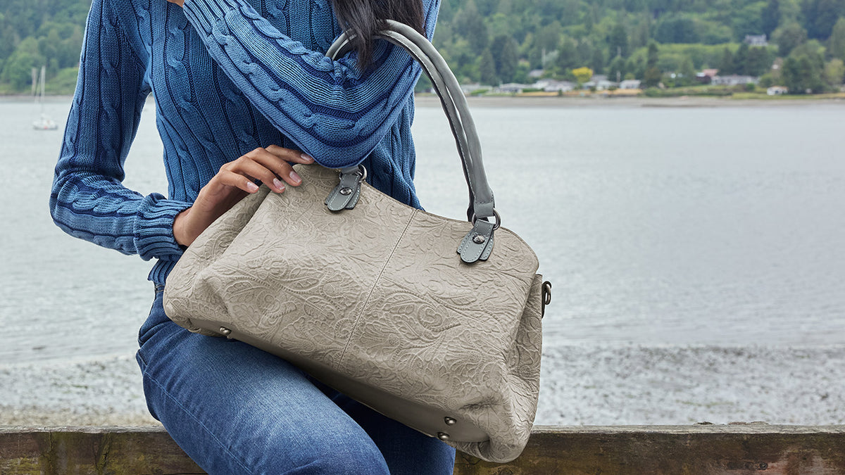 A woman in a blue cable-knit sweater and jeans sits by a lake holding a large textured taupe handbag with gray handles and hardware.