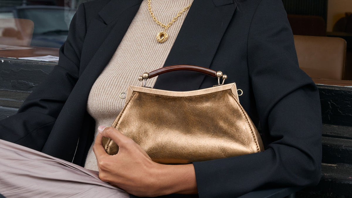 A person wearing a black blazer and beige knit top holds a metallic gold handbag with a brown top handle. The person is also wearing a gold necklace and ring, seated on a dark bench in an indoor setting.