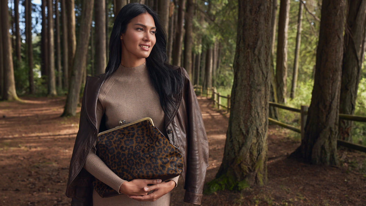 A woman standing in a forest holding a leopard print clutch bag.