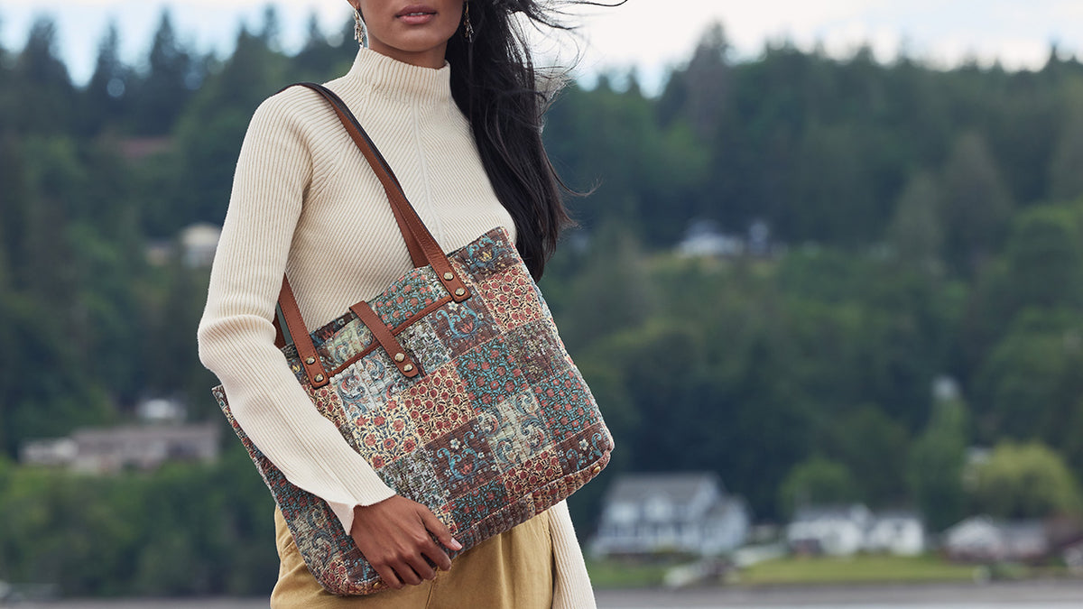 A woman wearing a cream ribbed turtleneck sweater and tan skirt carries a large patchwork tote bag with brown leather shoulder straps. The background is a soft-focus outdoor scene of a forested hillside.