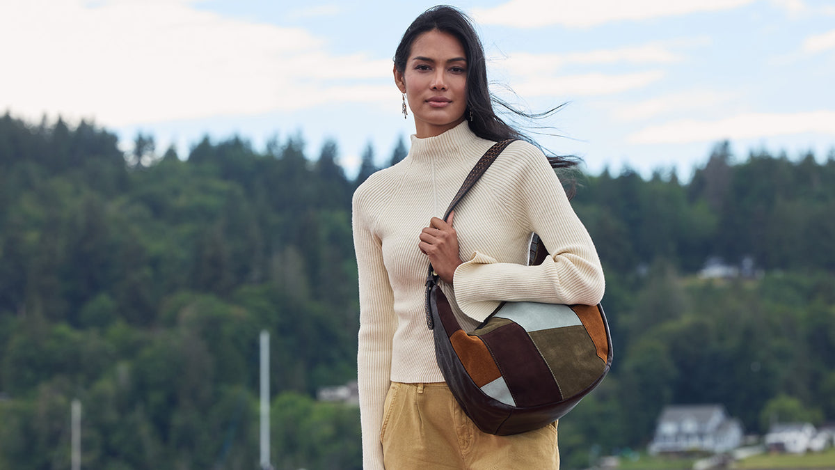 A woman wearing a cream ribbed turtleneck sweater and tan pants carries a brown patchwork shoulder bag with a dark strap, standing outdoors against a blurred background of green trees and a cloudy sky.