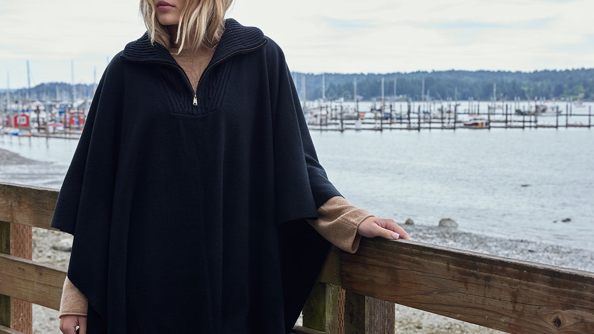 A woman wearing a black hooded poncho-style top with a half-zip neckline and tan sleeves, standing outdoors by a wooden railing with water and docks in the background.