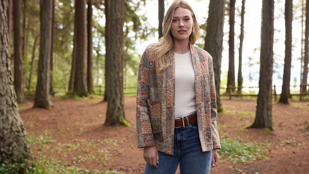 A woman stands in a forest wearing a multicolor patterned tweed blazer over a cream top and blue jeans.