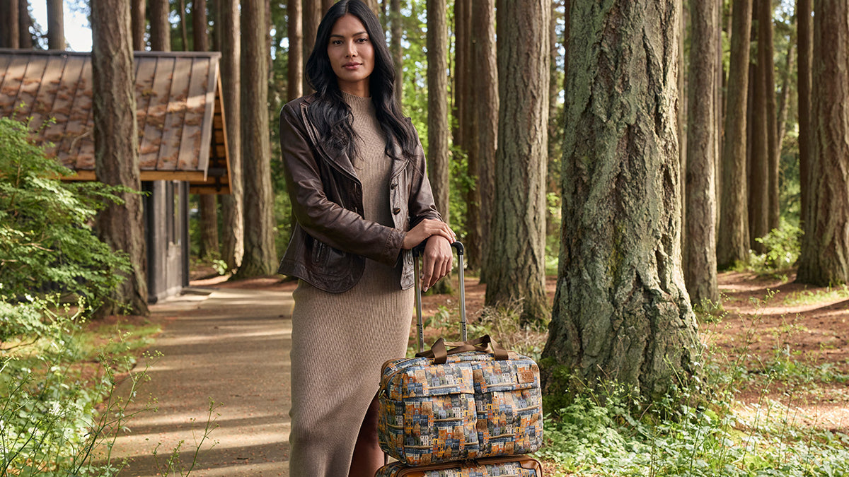 A woman stands in a wooded forest path next to a rustic cabin, wearing a brown leather jacket over a beige dress and holding a patterned blue and orange duffel bag.