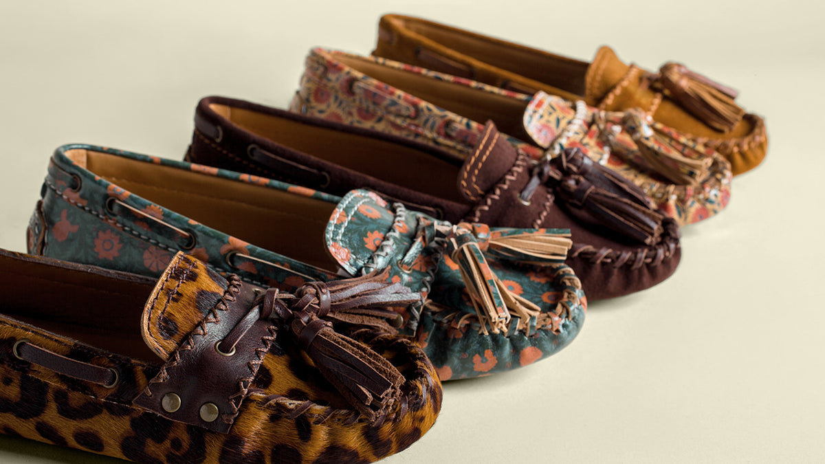 Four pairs of moccasin-style loafers arranged diagonally on a light beige background. The shoes feature diverse patterns including leopard print, teal floral, dark brown, and tan paisley, all with dark brown leather tassel straps.