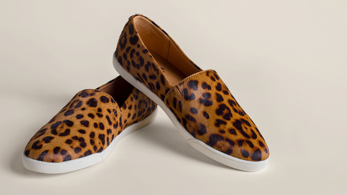 A pair of slip-on loafers with a brown and black leopard print pattern and white soles, displayed on a light beige background.