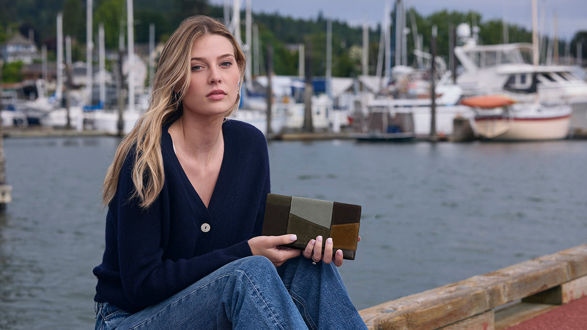 A woman sitting on a wooden dock by a harbor, wearing a navy blue cardigan and jeans while holding a rectangular geometric clutch in a dark brown and gold pattern. Boats and a marina are blurred in the background.