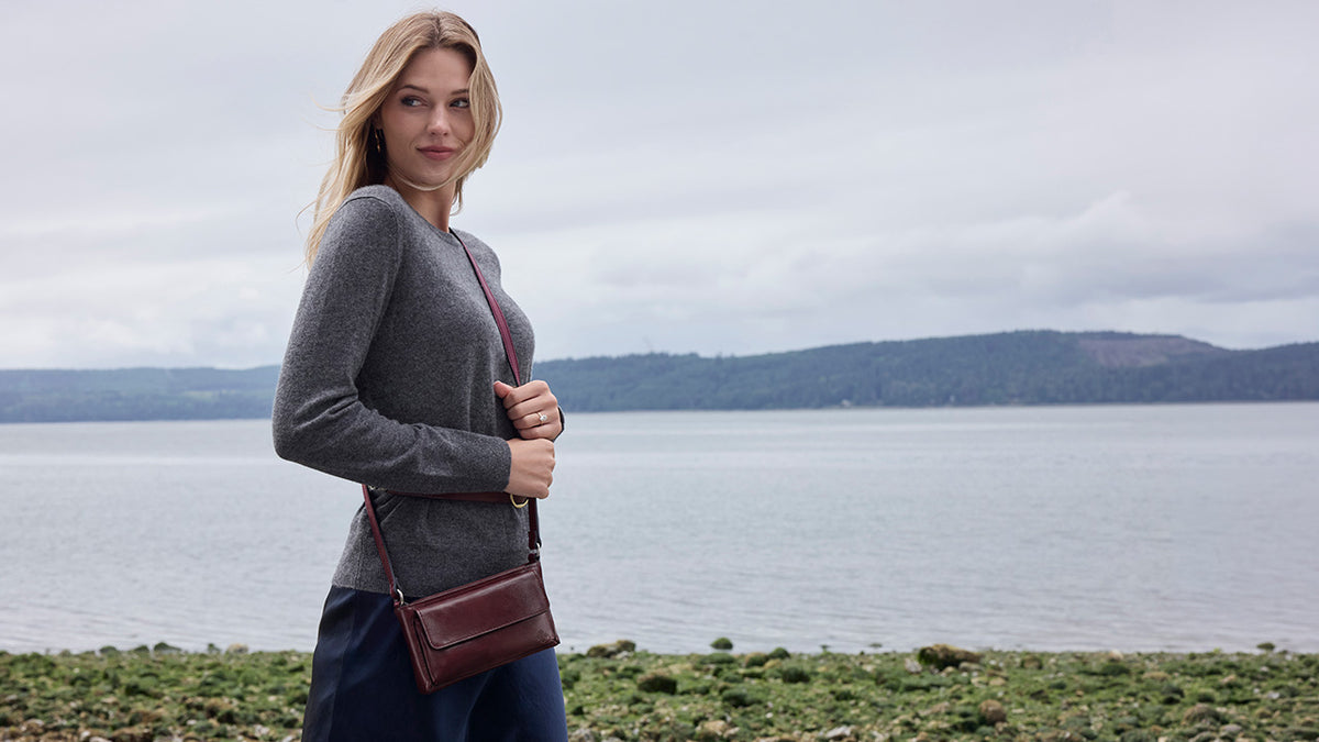A woman stands outdoors by the water wearing a gray long-sleeve top and carrying a dark brown leather crossbody bag with a thin strap.