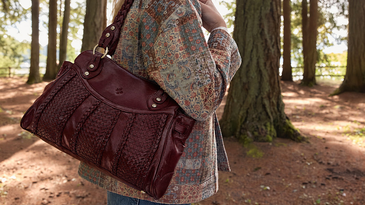 A person in a patterned cardigan carrying a large burgundy woven leather shoulder bag in a wooded outdoor setting.