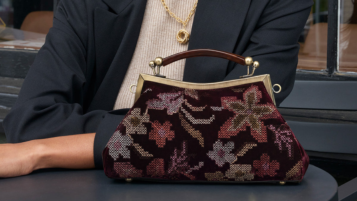A structured vintage-style clutch handbag with a dark burgundy velvet base and intricate floral embroidery in shades of pink, gold, and brown. The bag features a brown leather top handle with gold-tone metal rings and a gold-tone frame clasp.