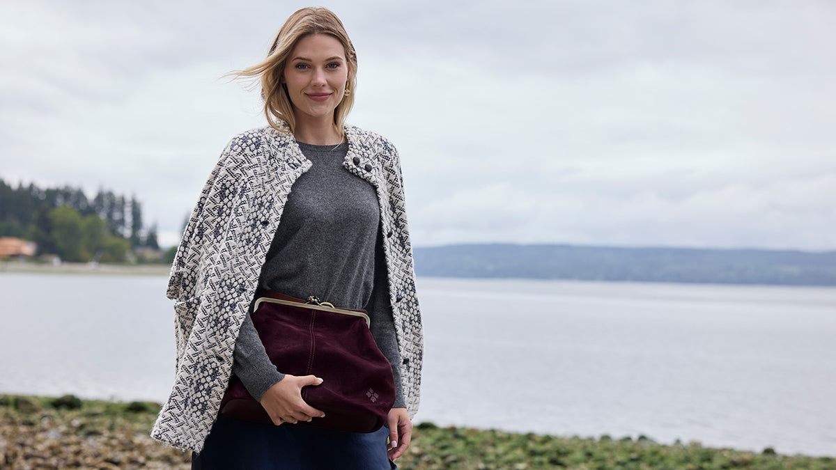 A woman standing outdoors by the water wearing a black and white patterned knit cardigan over a gray long-sleeve top, holding a burgundy clutch bag.