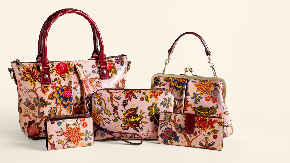 A coordinated handbag set on a warm off-white background, featuring a large tote bag, a medium shoulder bag, a wristlet clutch, and two small card cases. The bags have a pink floral and bird print with dark red leather handles and trim.