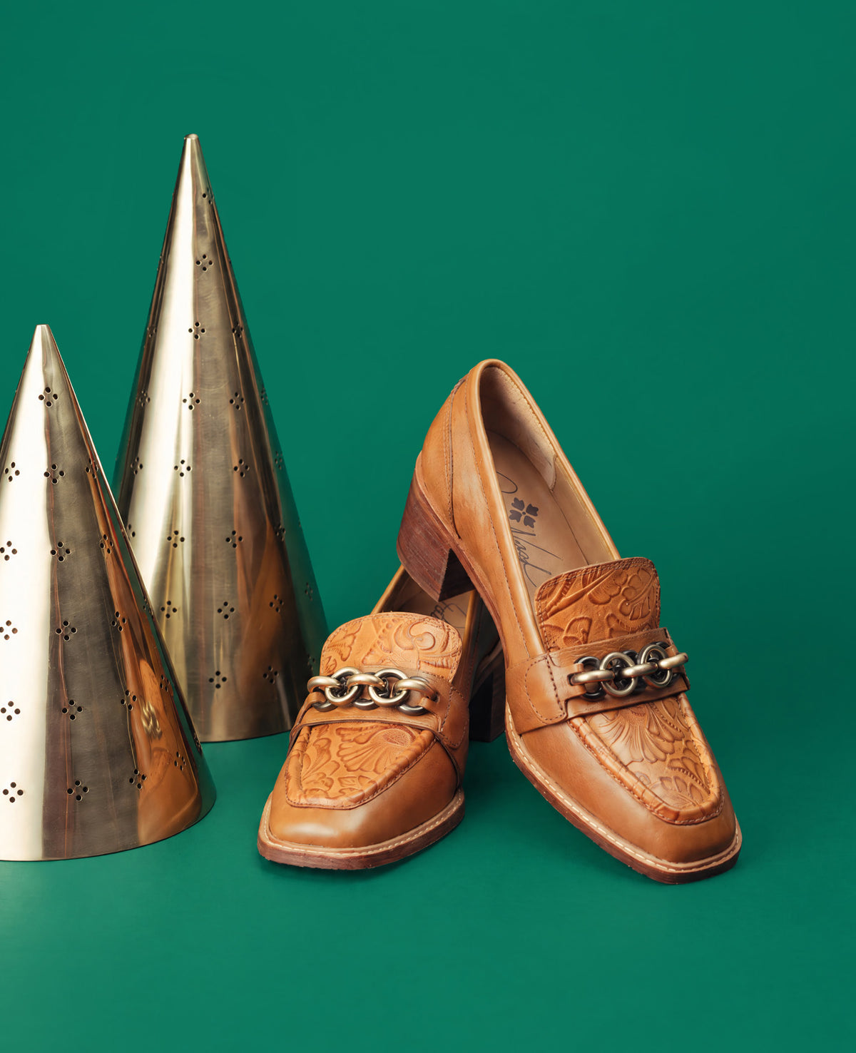 A pair of tan leather block-heel loafers with embossed floral detailing and a silver-tone chain link ornament across the vamp, displayed on a solid green background next to two metallic cone-shaped decorative objects.