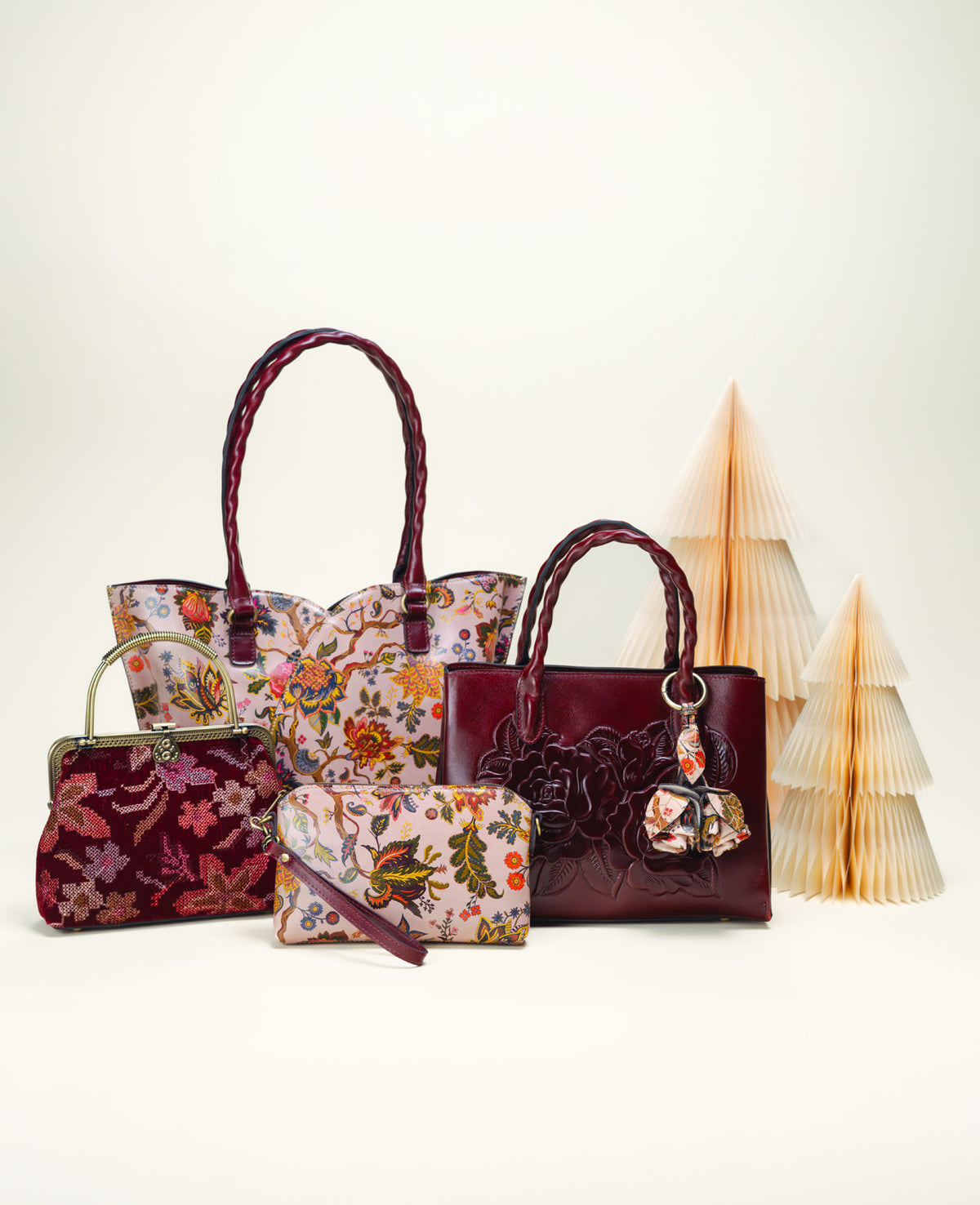 A coordinated handbag set displayed on a clean off-white studio background, including two floral tote bags, one embossed burgundy tote, one floral pouch, and one burgundy handbag with floral detail, arranged with decorative paper trees behind them.